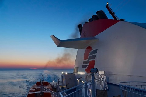 A ship leaves traces of smoke over the calm sea at dusk, New ferry port in southern Patras,