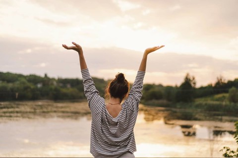 A woman raises her arms in joy while overlooking a peaceful lake at sunset. Her striped shirt and