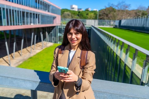 Young woman student smiling at camera while texting on a smartphone, standing on a walking bridge