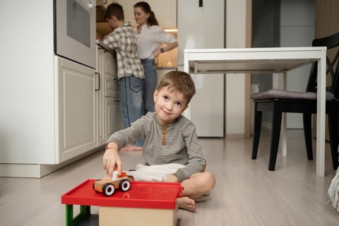 A young boy cheerfully plays with a toy car on the kitchen floor, while your family prepare food in