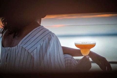 A woman sits on a terrace at sunset, savoring a cocktail in a serene atmosphere. The soft colors of