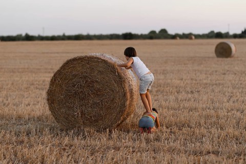 Two siblings interact with a large hay bale in a field, displaying curiosity and playfulness amidst