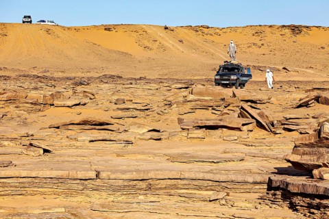 An off-road vehicle and people standing in front of sand dunes in a vast, rocky desert landscape,