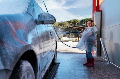 A joyful child in a clear raincoat and earmuffs sprays water on a foam covered car at a self serve