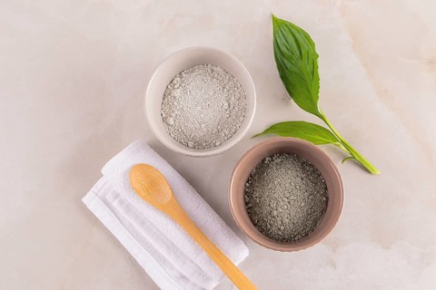 Top view of two bowls of cosmetic clay powder, a wooden spoon, and fresh leaves on a marbled