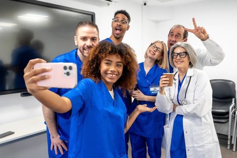 Diverse medical team in scrubs and lab coats smiling for a cheerful group selfie in a modern