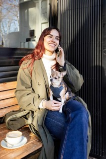 Smiling woman sits on a bench enjoying coffee with her small chihuahua in an outdoor cafe. She is