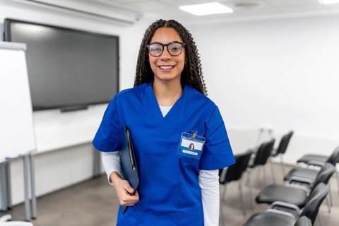 Young woman wearing a blue scrub and glasses, carrying a clipboard, smiling confidently while
