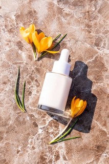 A white dropper bottle lies on a marble surface surrounded by bright yellow flowers. The minimalist