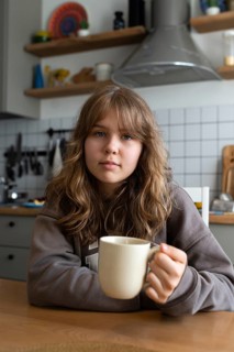 Teenage girl relaxing at home in a modern kitchen, holding a cup. Shelves with colorful decor and