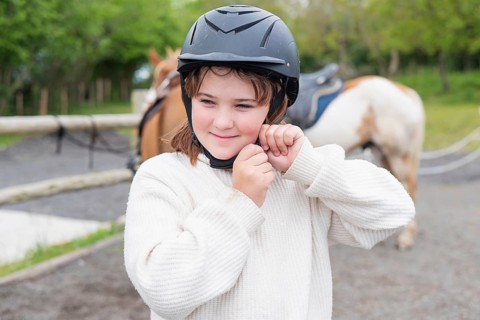 A young girl adjusts her helmet, preparing for a delightful horse ride on a sunny farm day. The
