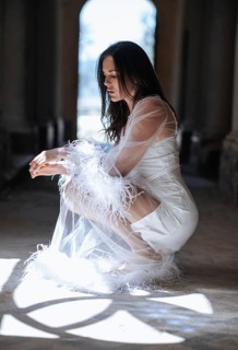 A bride in a short white gown with feather and sheer detailing kneels gracefully in a sunlit