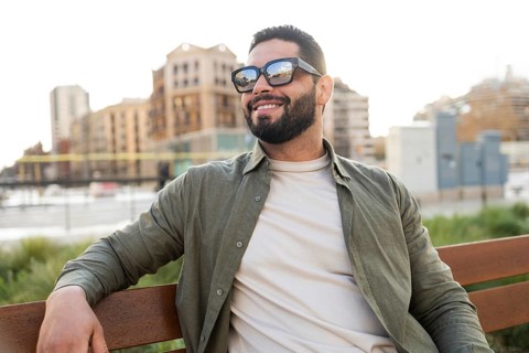 A stylish man wearing sunglasses and a casual outfit relaxes on a bench, smiling while gazing at an