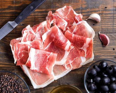 Thinly sliced prosciutto arranged on a wooden board, accompanied by black olives and a bowl of