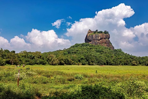 A massive rock rises above a wide, green field under a partly cloudy sky, The Lion Rock near