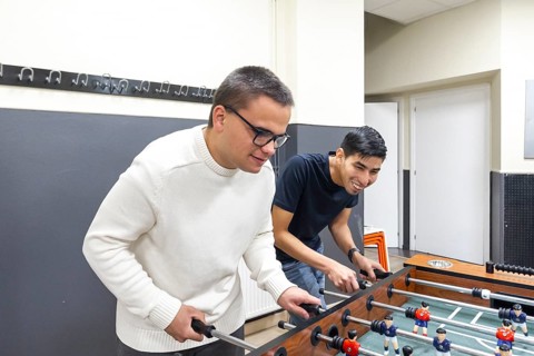 Two friends play foosball at a mental health leisure center, smiling and engaging in friendly