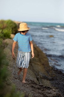 A young boy with a straw hat and striped clothing walks carefully along a rocky beach path beside