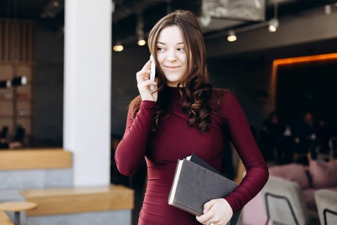 A young female student stands indoors, balancing books while talking on her phone. Her expression