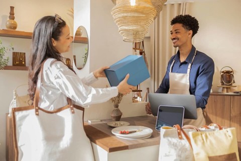 A customer in a stylish bag store hands a gift box to a friendly shop assistant. The setting is