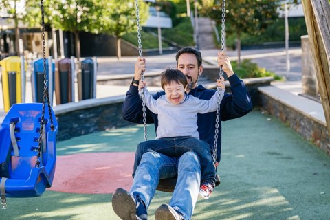 A father and his son with Down syndrome share a joyful moment on a swing in a sunny park. The child