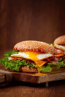 Two juicy burgers sit on a wooden board, each filled with lettuce, bacon, cheese, and fried eggs