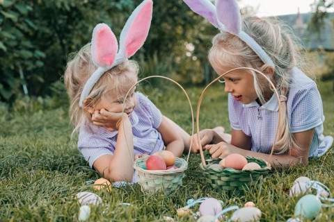 Two girls wearing bunny ears enjoy an Easter egg hunt in a lush garden. They sit among pastel