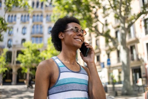 Smiling non-binary individual with afro hair enjoys a sunny day while talking on the phone, wearing