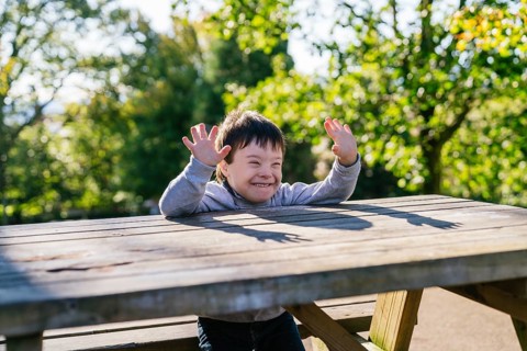 A cheerful boy with Down syndrome raises hands playfully over a wooden picnic table in a sunlit