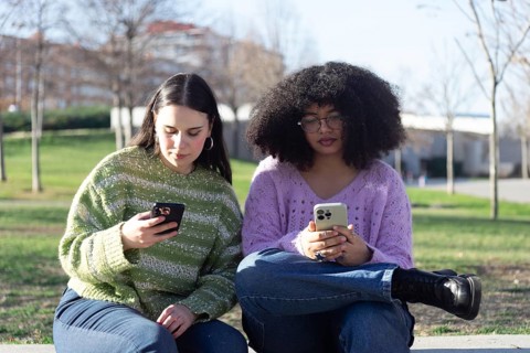 Two women sit on a bench in a park, each focused on their smartphone. They are dressed in casual