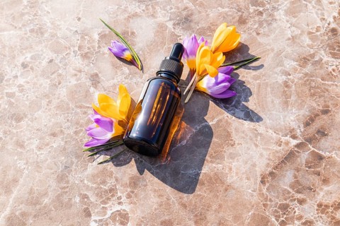 A brown essential oil bottle for skincare lies on a marble surface, surrounded by vibrant crocus