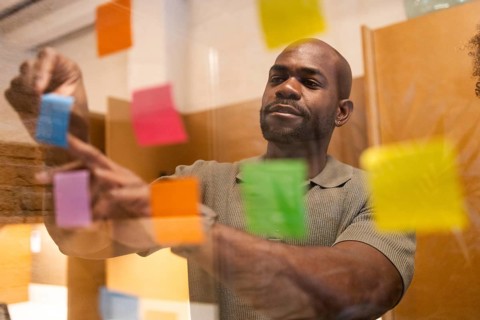 A professional in a coworking space in Barcelona places vibrant post-it notes on a glass wall. The