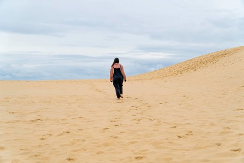 A solitary woman walks across expansive desert sand under a cloudy sky. The vast landscape and