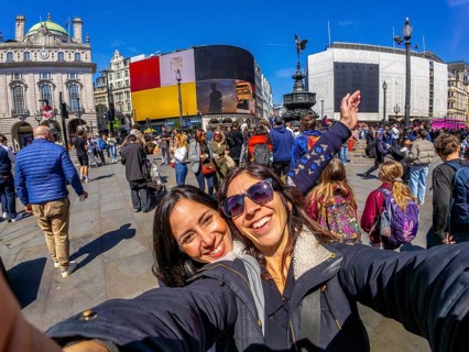 Two happy tourists taking a selfie with fisheye lens in piccadilly circus, london, with crowds of