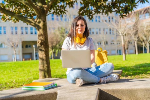 Portrait of a joyful student using laptop sitting on the ground outside the campus