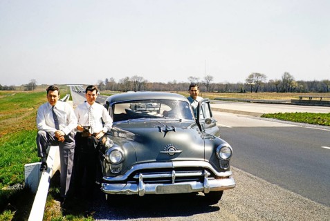 Three men in business suits standing next to an Oldsmobile 88 on a motorway, USA around 1953