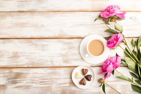 Cup of cioffee with chocolate candies, pink peony flowers on white wooden background. top view,