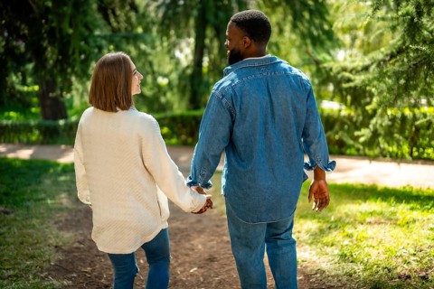 Rear view of a multi-ethnic couple walking holding hands along a park