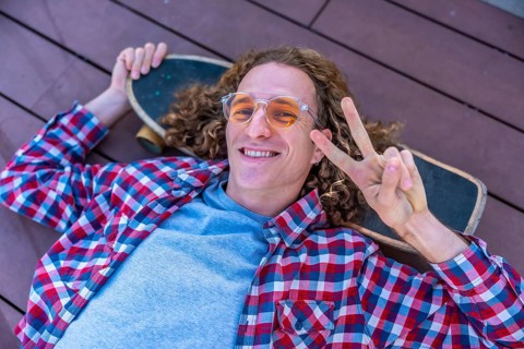 Smiling young man with curly hair relaxing on a skateboard, flashing a peace sign while lying on a