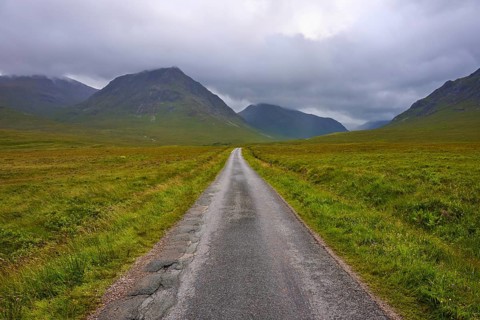 Endless straight road through vast grassland, framed by mountains under a cloudy sky, Glen Etive,