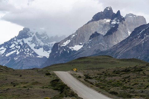 Gravel road with yellow curve signs leads through steppe landscape to the Cuernos del Paine