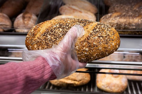 A hand in a pink sleeve uses a plastic glove to pick up a fresh multigrain loaf from a bakery