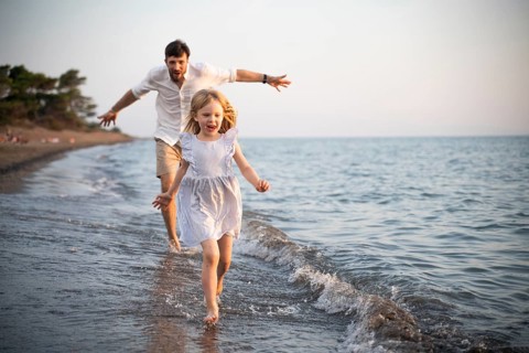 A father and his daughter share a joyful moment running along the shoreline of a beach in Italy.