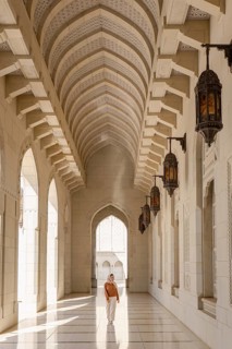 A woman in modest attire walks through the serene, sunlit corridors of the Sultan Qaboos Mosque in
