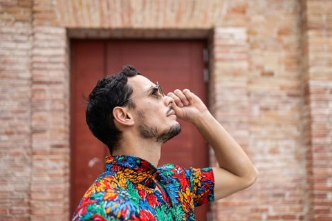 A man in a vibrant, floral shirt stands confidently against a brick wall. The colorful attire and