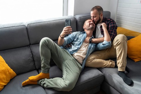 A gay couple relaxes on a sofa, taking a selfie. One partner lies on the other's lap, showcasing a