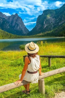 Woman wearing summer hat sitting on a wooden fence, admiring lake dobbiaco and the dolomites, italy