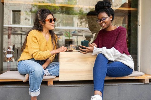 Two friends sit together outside a cozy cafe, enjoying coffee and sharing a phone. They wear casual