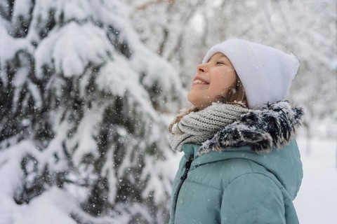 A joyful child in winter attire smiles while enjoying falling snow. The snowy landscape and