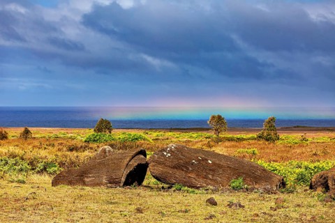 Large boulders in a meadow with a rainbow over the sea in the background, the Moai statues on
