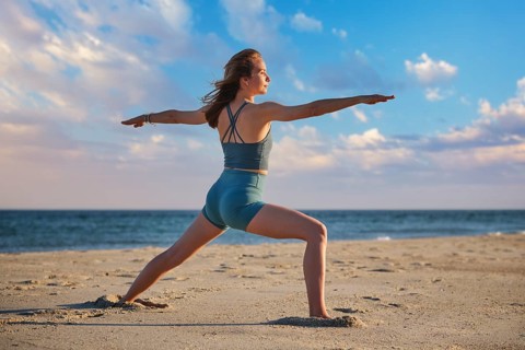 A woman performs Virabhadrasana 2, or Warrior 2 pose, on a beach during sunset, enjoying the